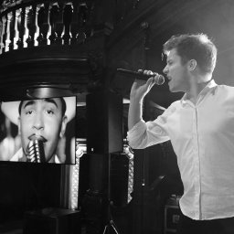 FOKUS GROUP Michał Margas - Czarno-białe zdjęcie wokalisty z mikrofonem na scenie, obok ekran z wizerunkiem innego artysty. W tle balustrada i oświetlenie sceniczne.