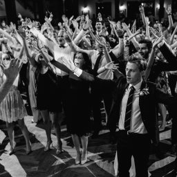 FOKUS GROUP Michał Margas - Czarno-białe zdjęcie z parkietu tanecznego, gdzie tłum ludzi w eleganckich strojach unosi ręce w górę, wyrażając radość podczas imprezy. W centrum widać mężczyznę w garniturze.