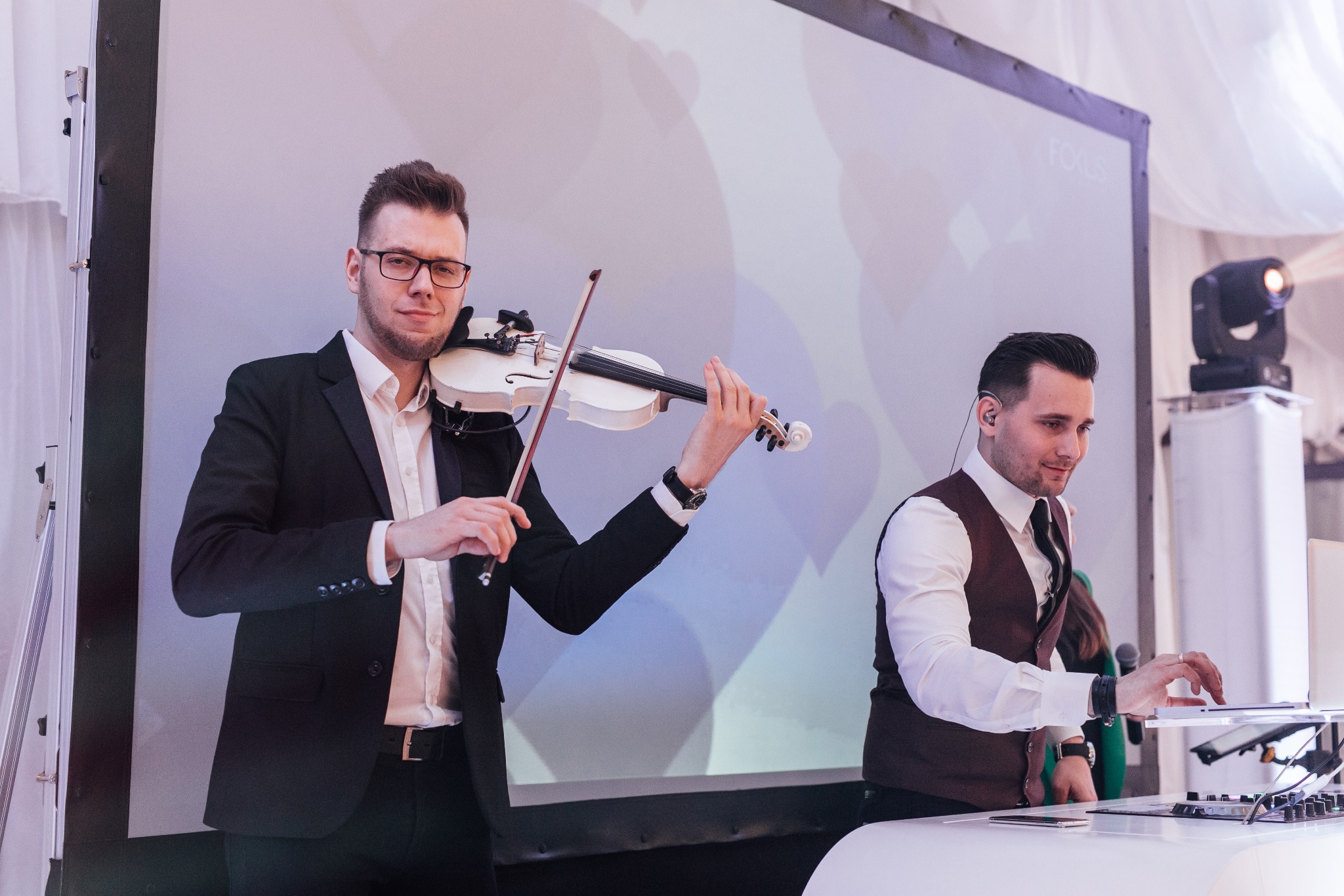 Duet muzyczny: skrzypek w czarnym garniturze gra na białych skrzypcach, obok DJ w kamizelce obsługuje sprzęt. W tle ekran i oświetlenie sceniczne.