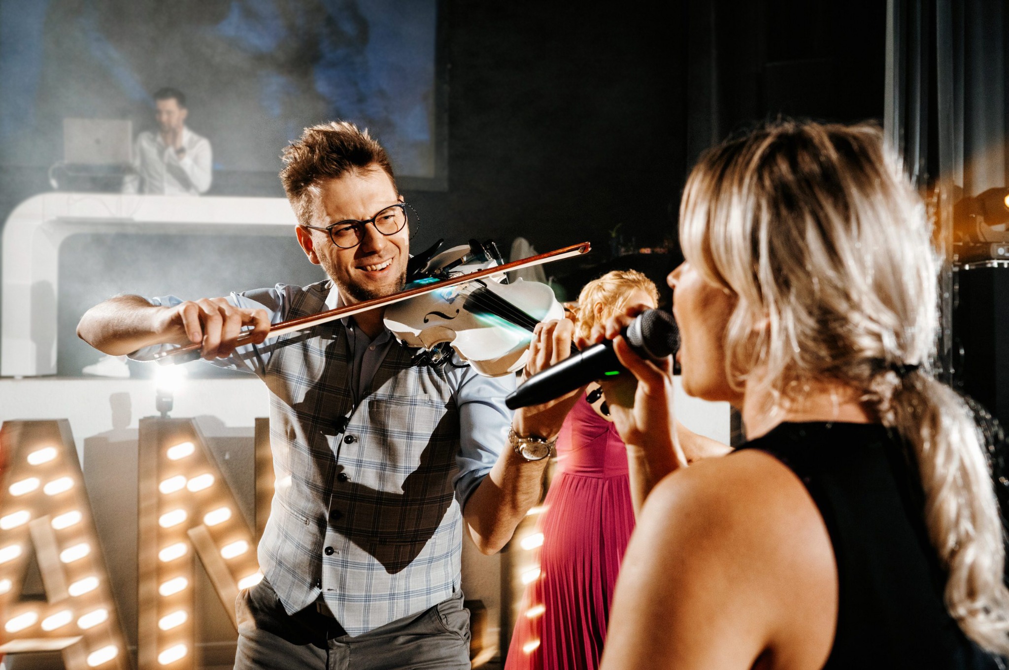 Uśmiechnięty skrzypek gra na białych skrzypcach, obok wokalistka z mikrofonem. W tle DJ za konsoletą i dekoracyjne litery z żarówkami. Scena w trakcie występu.