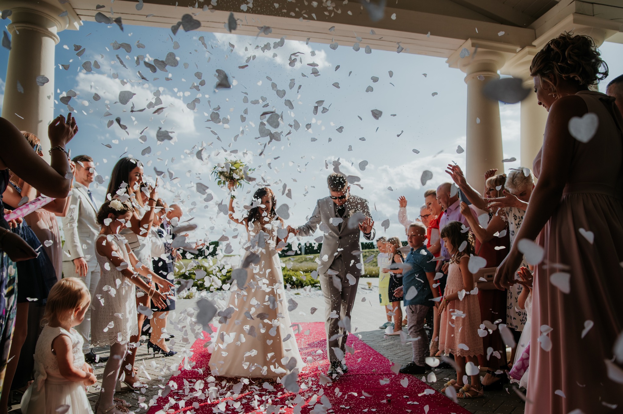Para młoda w szarych garniturach i białej sukni wychodzi spod kolumnady w deszczu konfetti w kształcie serc, otoczona gośćmi weselnymi.