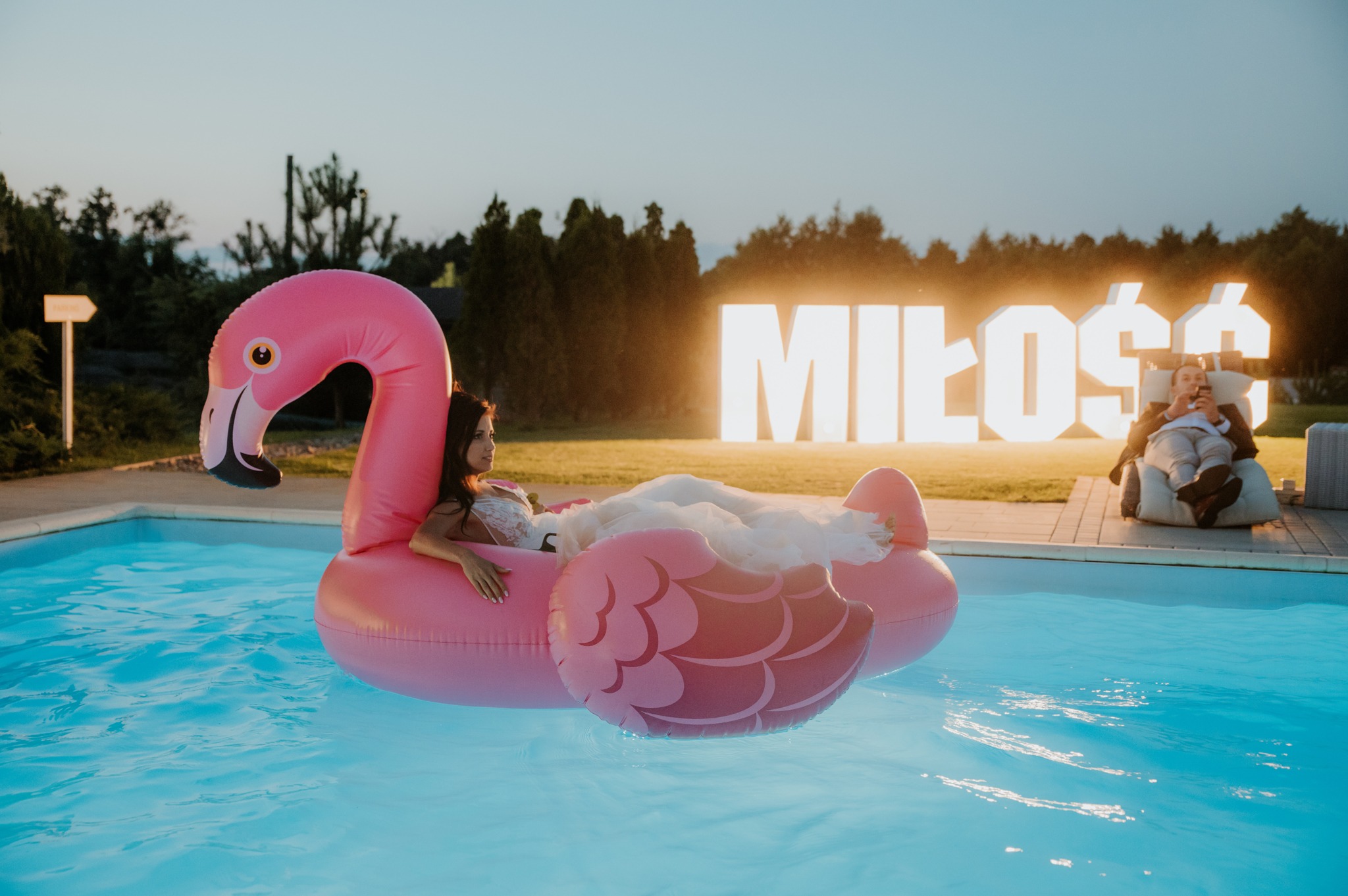 Panna młoda w sukni na różowym flamingowym materacu w basenie, w tle podświetlony napis 'Miłość' i mężczyzna w fotelu. Romantyczna sceneria ślubna.