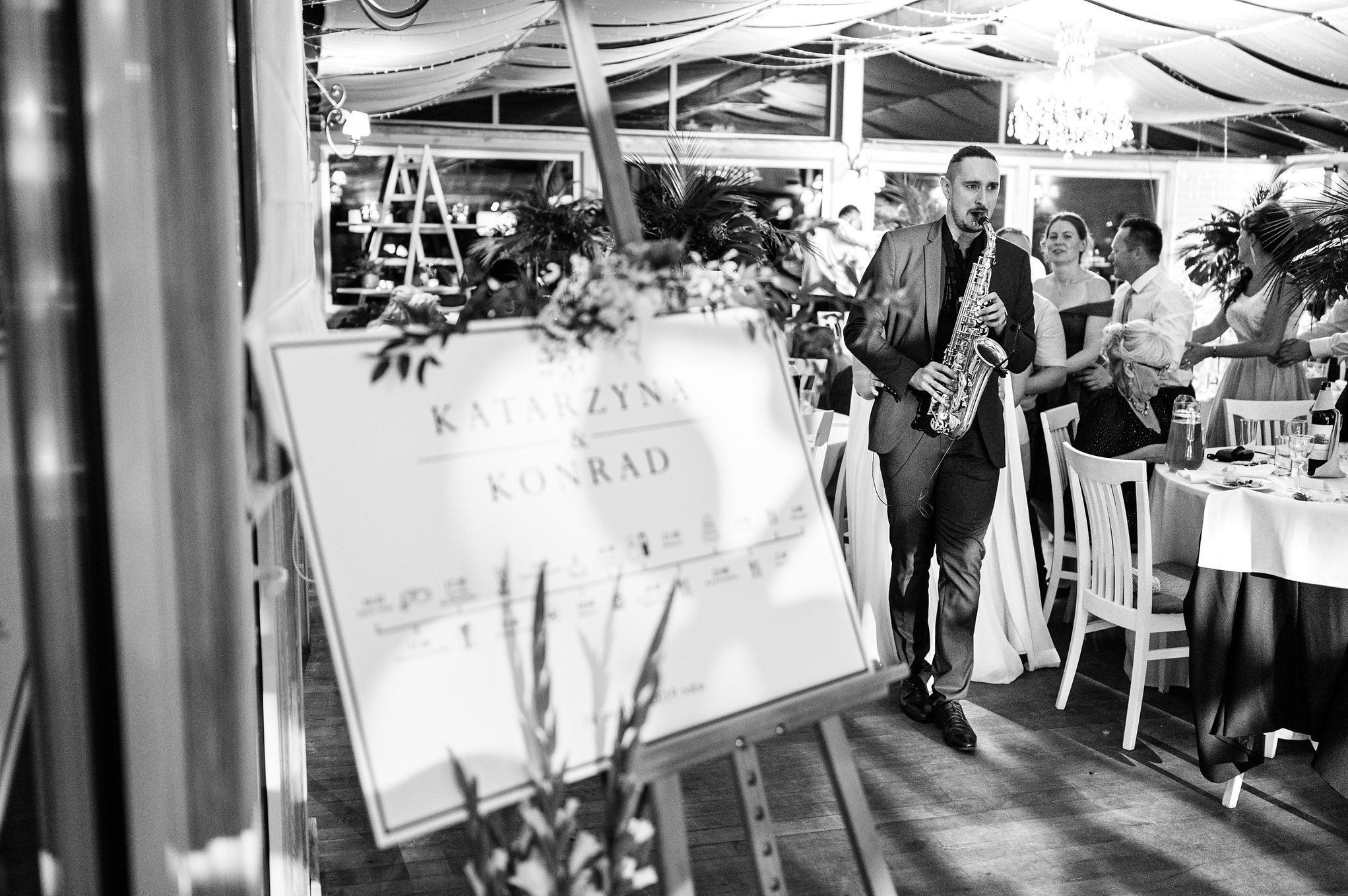 Czarno-białe zdjęcie saksofonisty w garniturze grającego na saksofonie na weselu, z tablicą powitalną z imionami pary młodej na pierwszym planie.