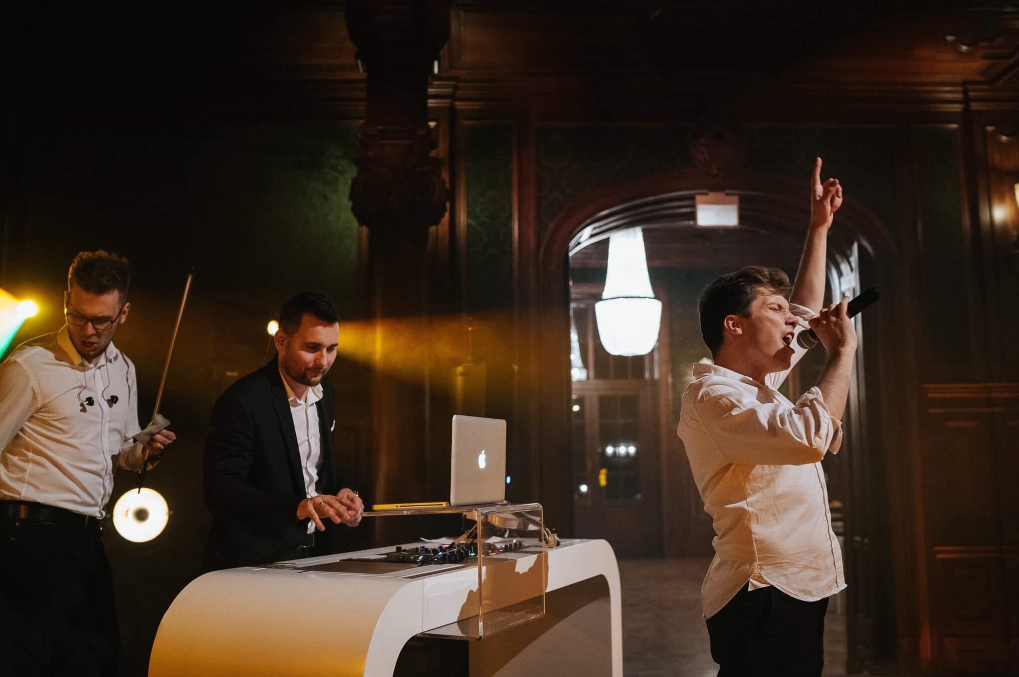 Zespół muzyczny na scenie: wokalista z mikrofonem, skrzypek i DJ z laptopem. W tle dekoracyjne, ciemne ściany i oświetlenie sceniczne.