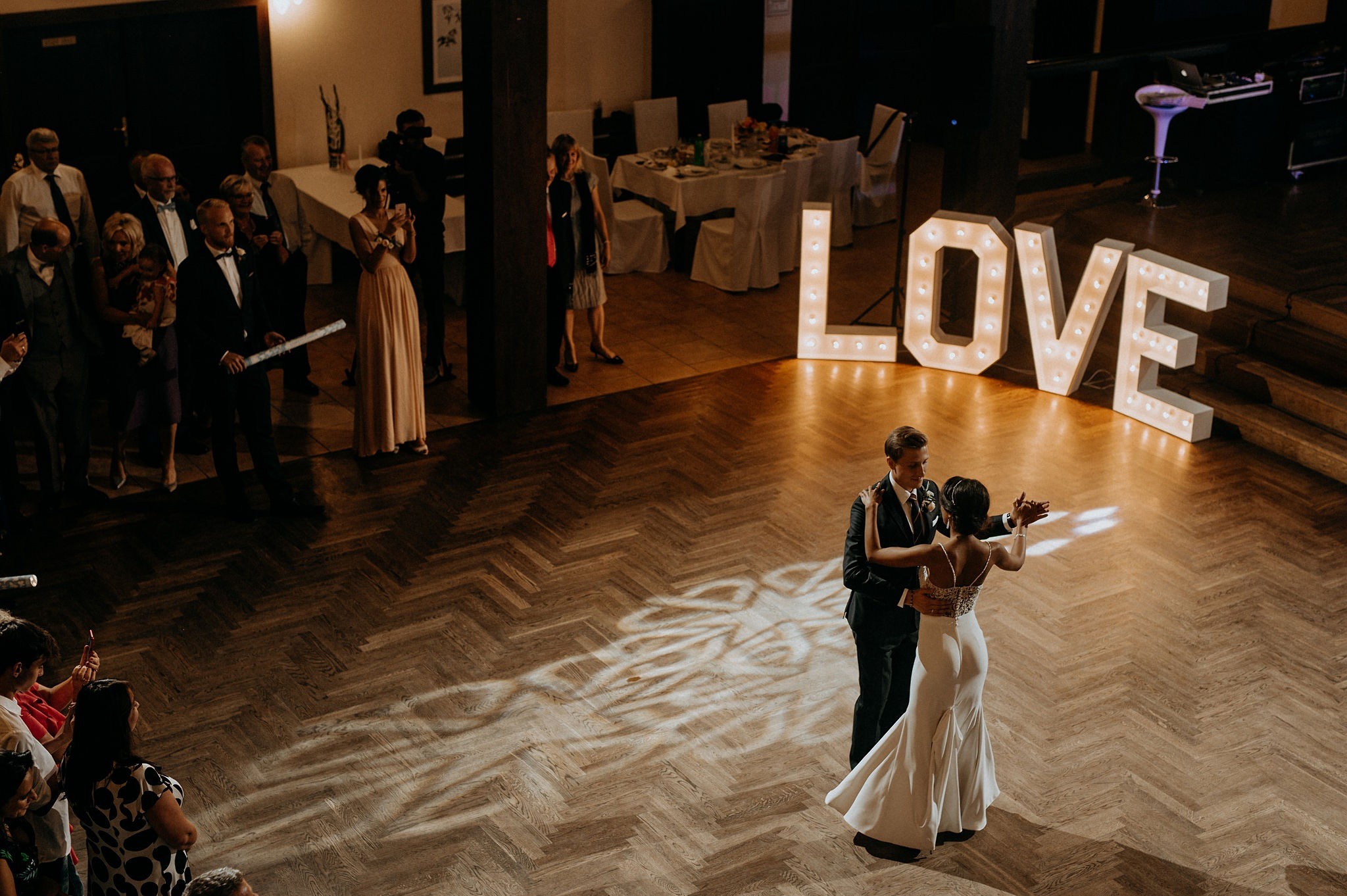Para młoda tańczy na parkiecie w sali weselnej, oświetlona dekoracyjnym napisem LOVE. Goście obserwują, w tle stoły i kolumny.