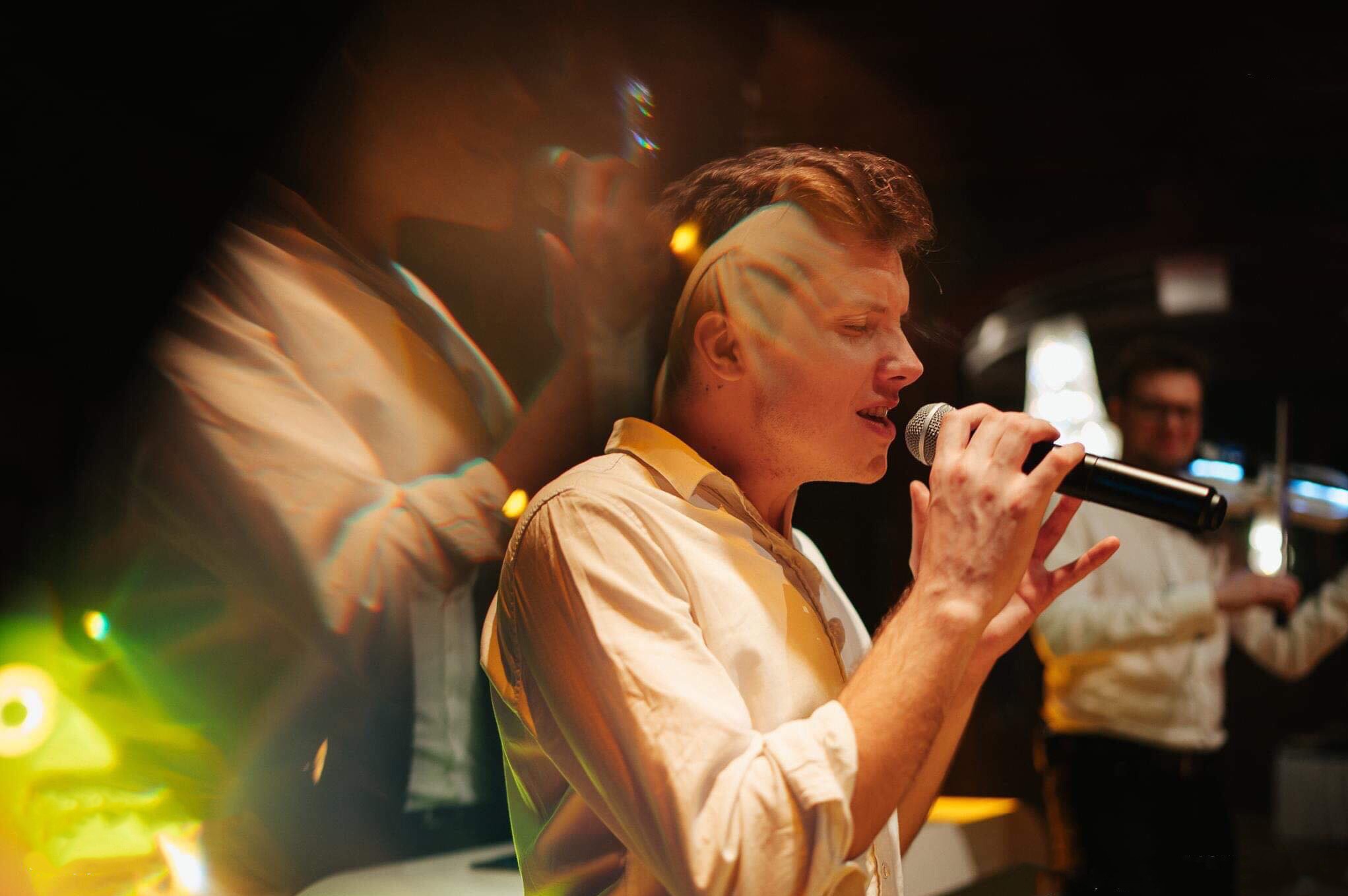 Wokalista z mikrofonem w dłoni, w tle muzyk z skrzypcami, efekt rozmycia światła. Ujęcie z występu na żywo, oświetlenie sceniczne.