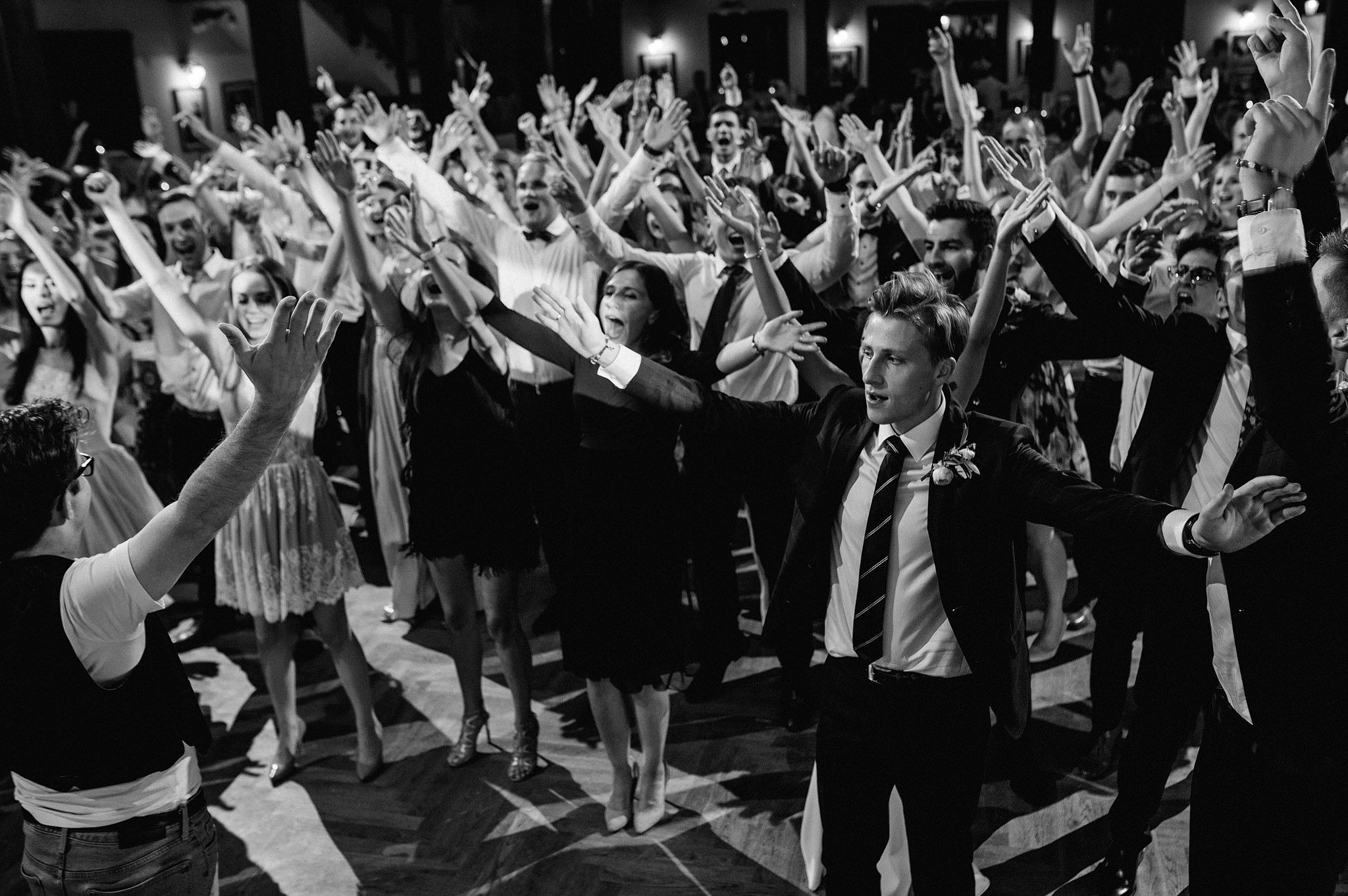 Czarno-białe zdjęcie z parkietu tanecznego, gdzie tłum ludzi w eleganckich strojach unosi ręce w górę, wyrażając radość podczas imprezy. W centrum widać mężczyznę w garniturze.