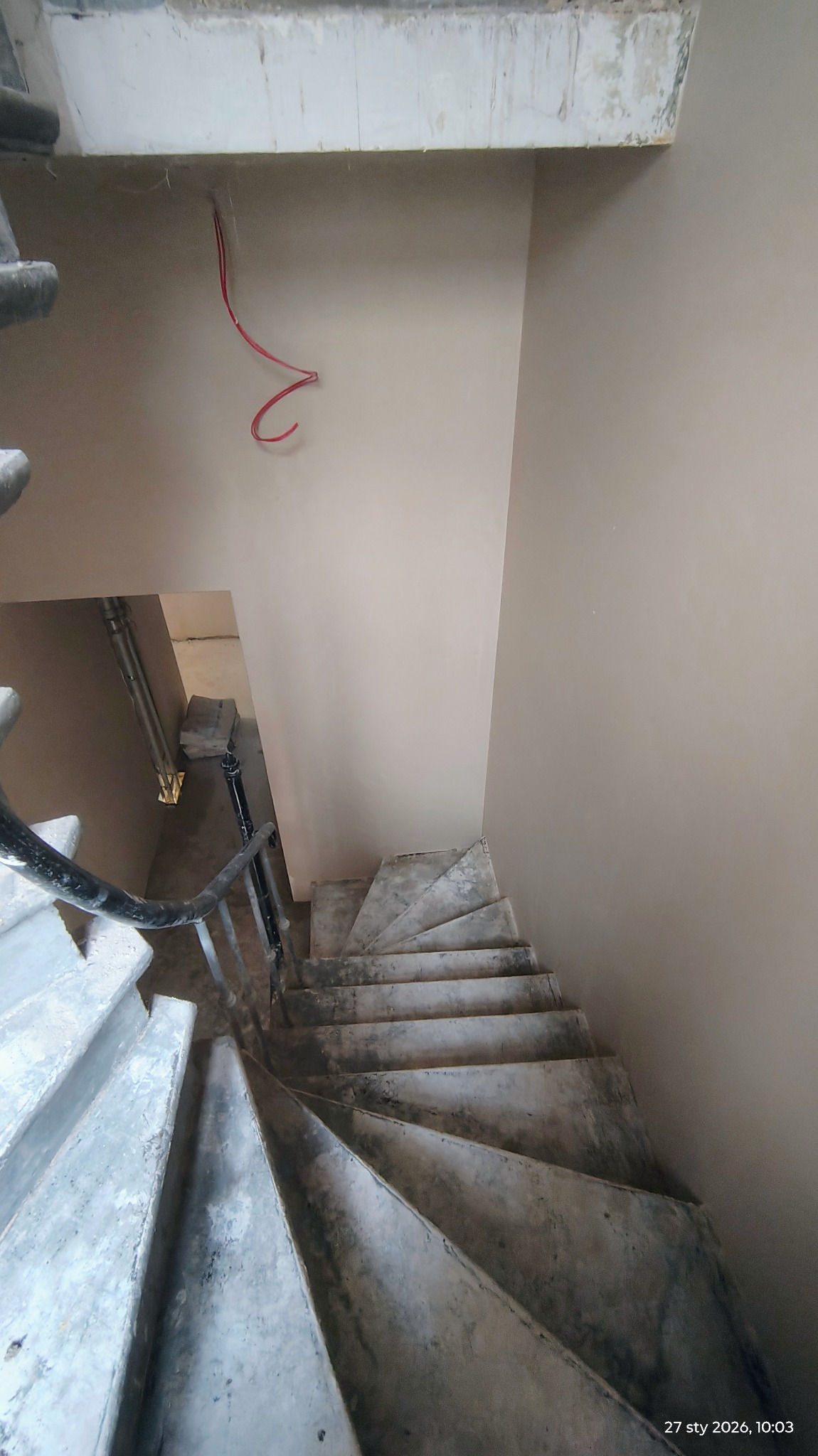 Klatka schodowa po remoncie. Ściany pokryte gładzią w odcieniach beżu, widoczne schody z szarego betonu. U góry wystające kable. Ujęcie z góry.