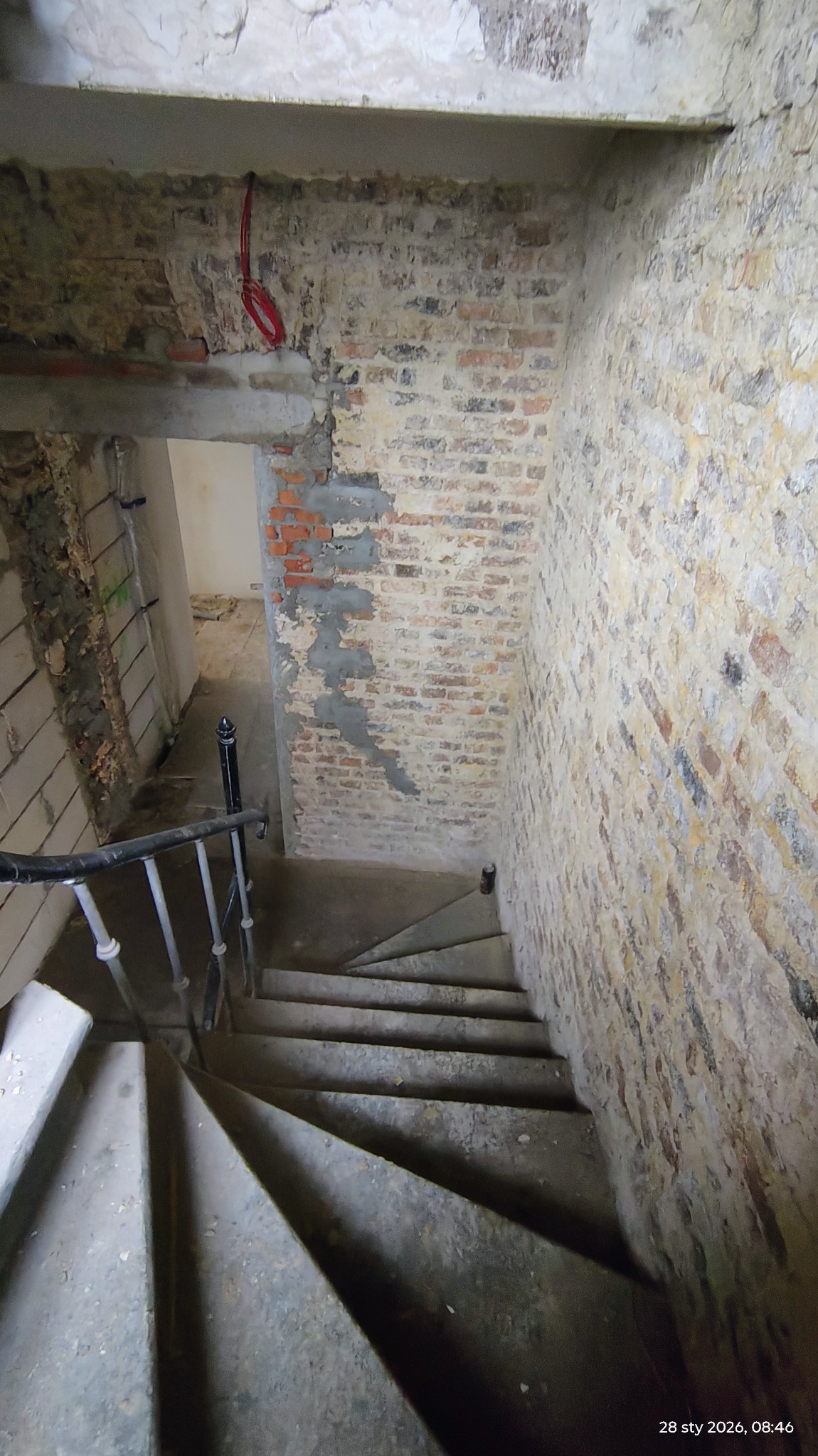Klatka schodowa w trakcie remontu: surowe cegły, częściowo otynkowane ściany, widoczne przewody elektryczne. Ujęcie z góry na schody.
