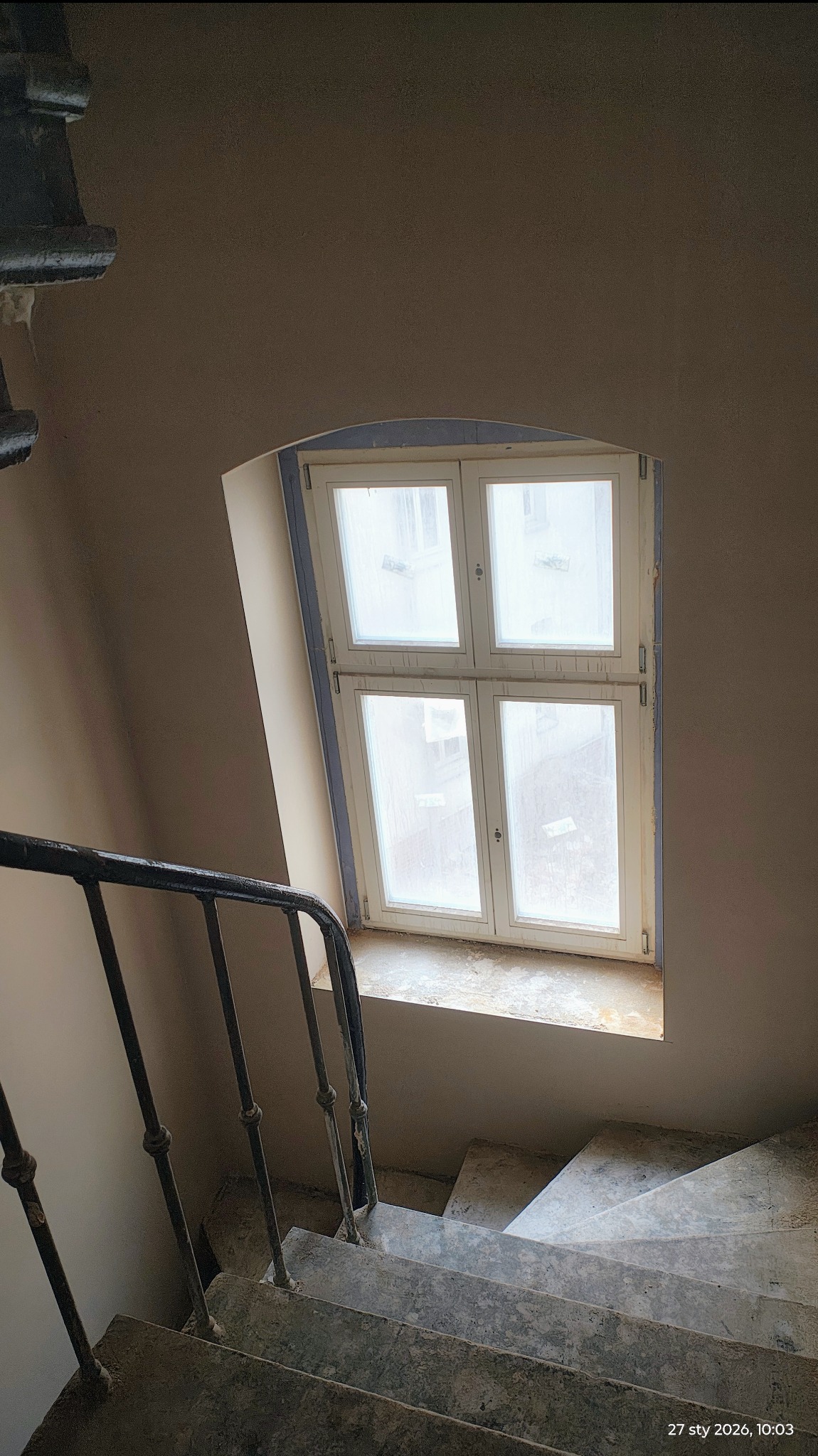 Klatka schodowa w trakcie remontu: ściany po nałożeniu gładzi, okno zabezpieczone folią, widoczne schody i metalowa balustrada. Ujęcie z góry.