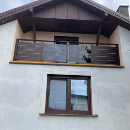 Usługi Ślusarsko Spawalnicze Wiktor Ozdowy - Balkon z brązową balustradą, częściowo szklaną, na tle szarej elewacji domu. Widoczne poziome deski i odbicie drzewa w szkle. Ujęcie z dołu, pochmurne niebo.