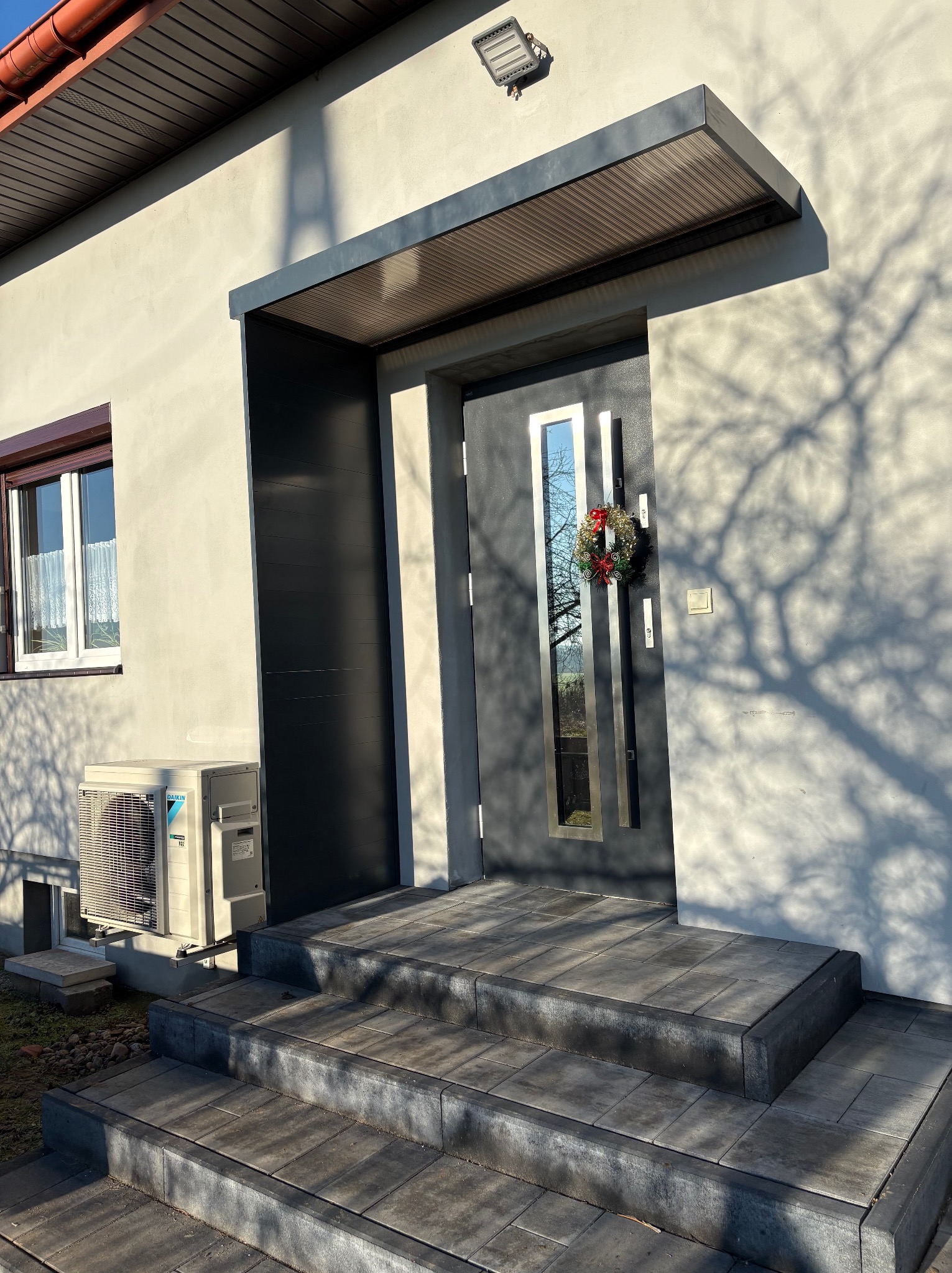 Nowoczesne wejście do domu z szarymi schodami i zadaszeniem. Drzwi z metalowymi elementami i świątecznym wieńcem. Cień drzewa na elewacji.