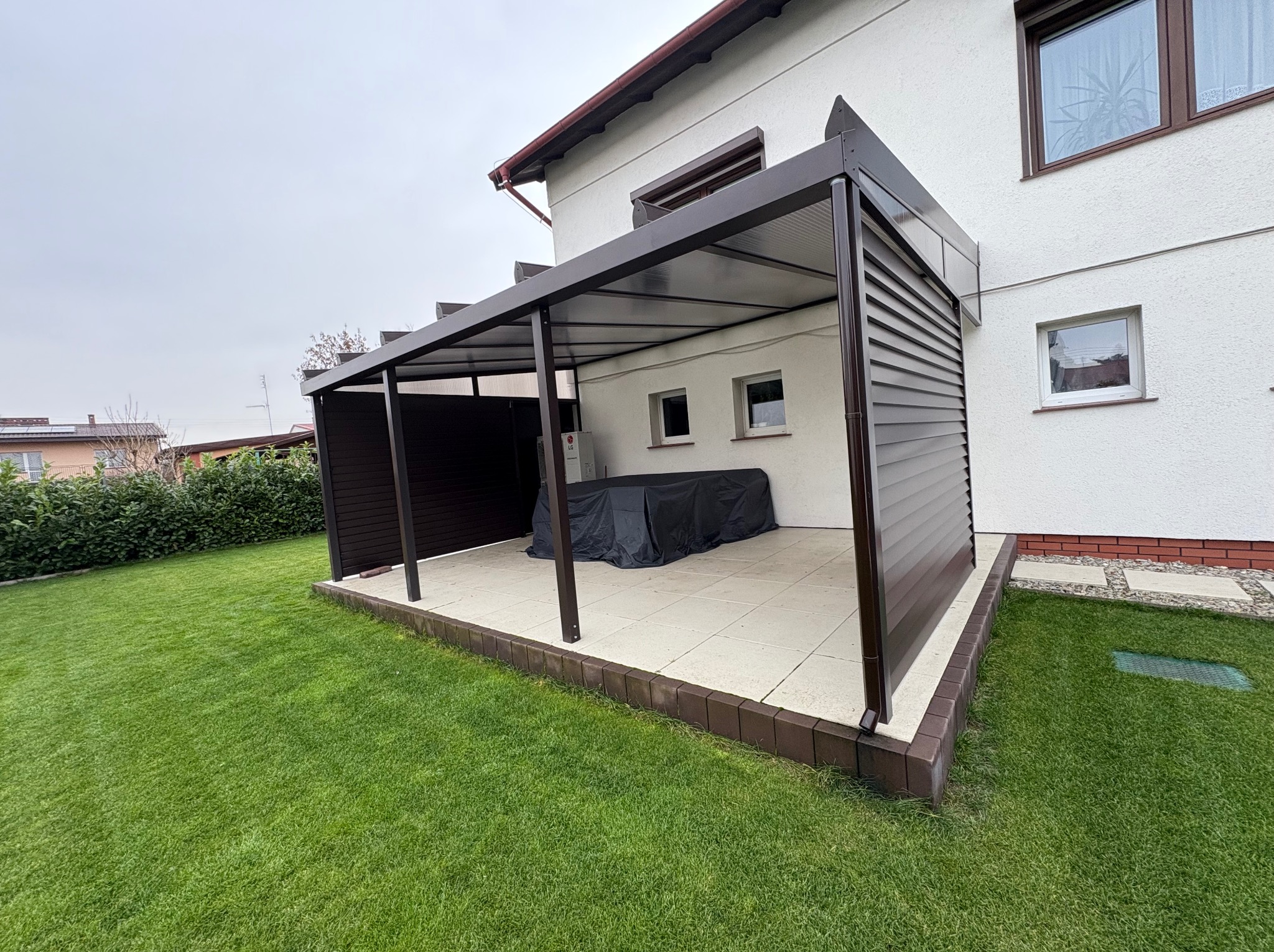 Nowoczesna, brązowa pergola z częściową zabudową ścianką lamelową, przylegająca do białego domu. Widok z zewnątrz, na pierwszym planie trawnik.