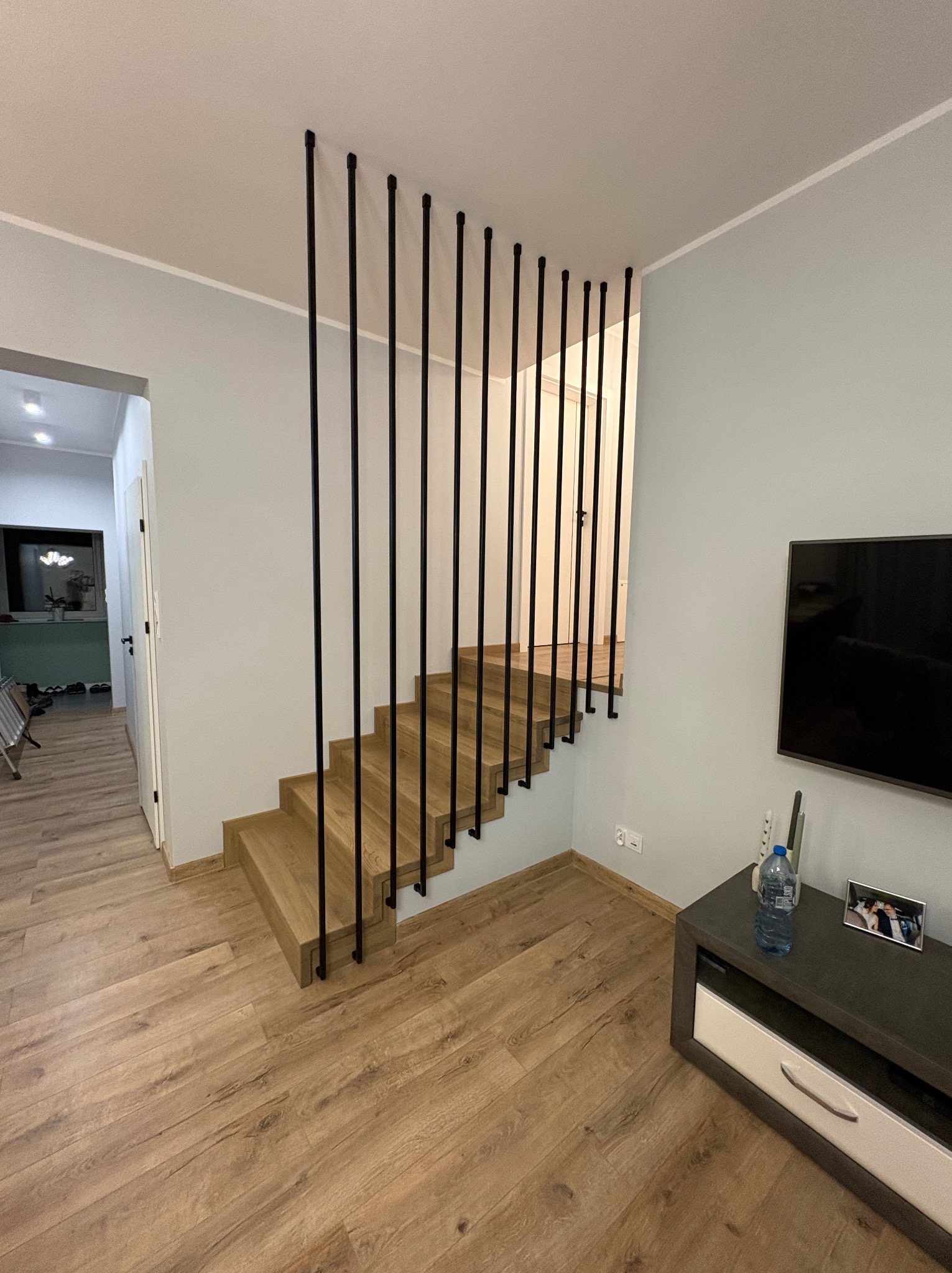 Drewniane schody z czarnymi, pionowymi elementami balustrady, widoczne z boku w jasnym wnętrzu. Minimalistyczny design, naturalne światło, parkiet na podłodze.