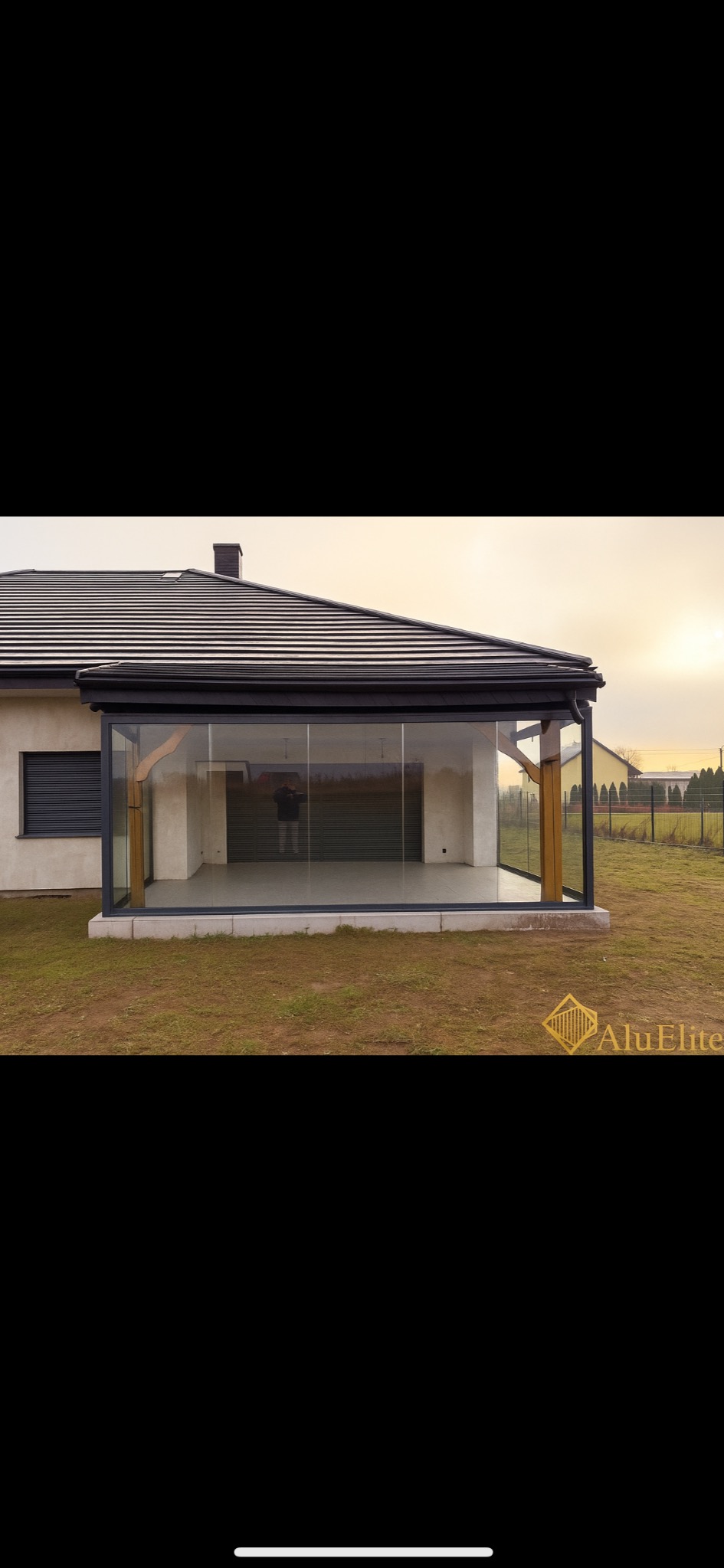 Szklana zabudowa tarasu z drewnianymi elementami konstrukcyjnymi, widoczna z zewnątrz, na tle domu jednorodzinnego i ogrodu. Minimalistyczny design.