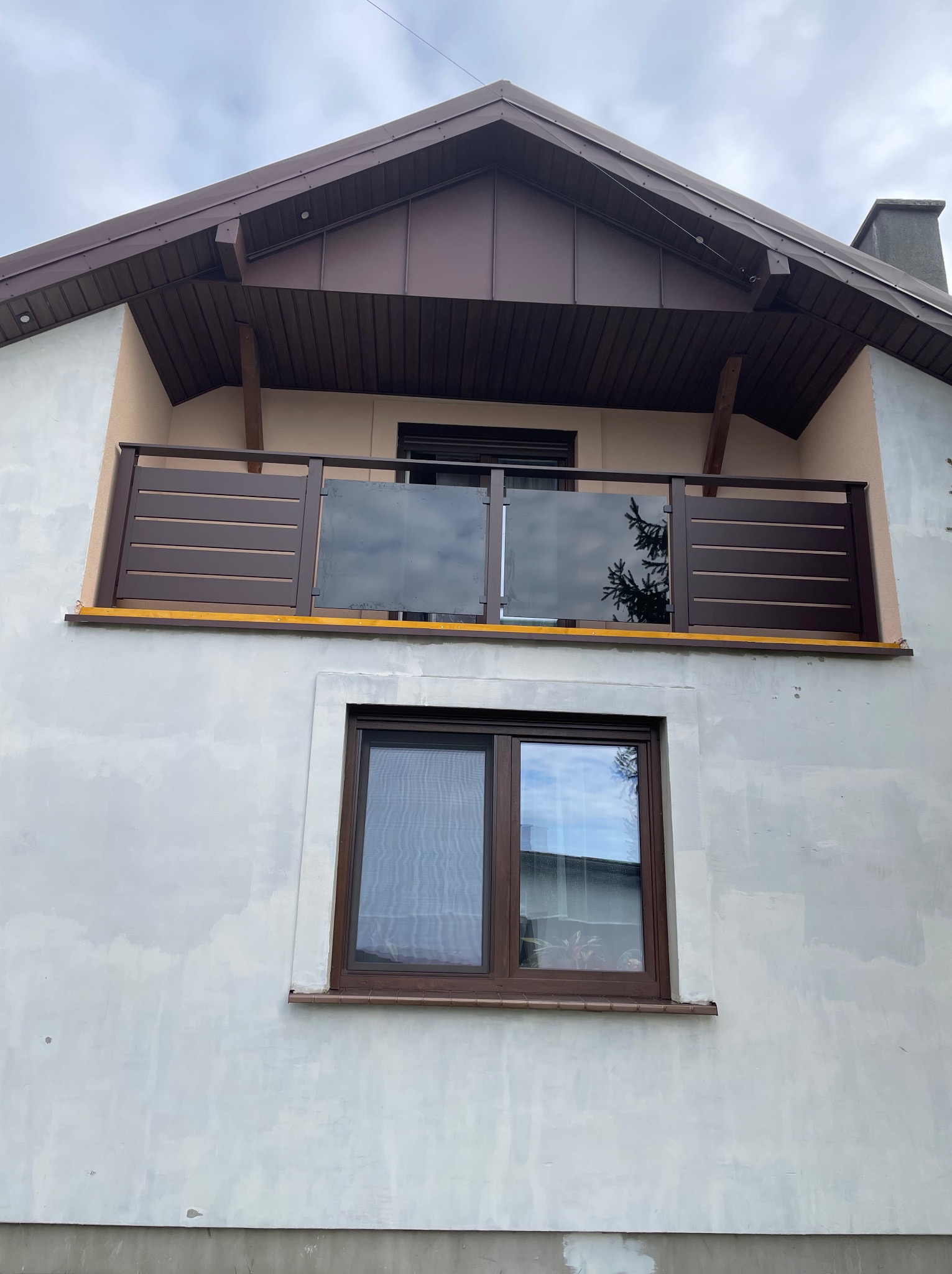 Balkon z brązową balustradą, częściowo szklaną, na tle szarej elewacji domu. Widoczne poziome deski i odbicie drzewa w szkle. Ujęcie z dołu, pochmurne niebo.