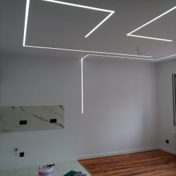 Usługi budowlane brukarstwo Marek Jacyno-Onuszkiewicz - Nowoczesne oświetlenie LED w kształcie geometrycznym na suficie, białe ściany, marmurowe płytki podłogowe i drewniana podłoga. Widoczny fragment ściany z płytkami.