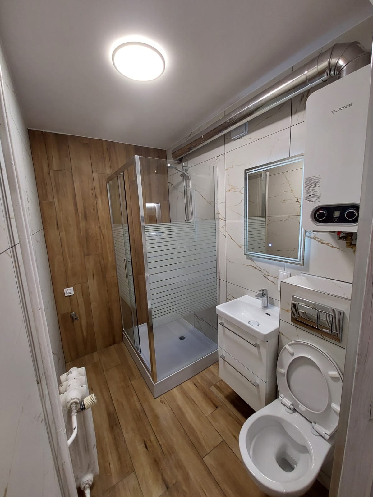 Nowoczesna łazienka po remoncie: kabina prysznicowa, umywalka z szafką, toaleta w zabudowie i marmurowe płytki z drewnianymi akcentami.