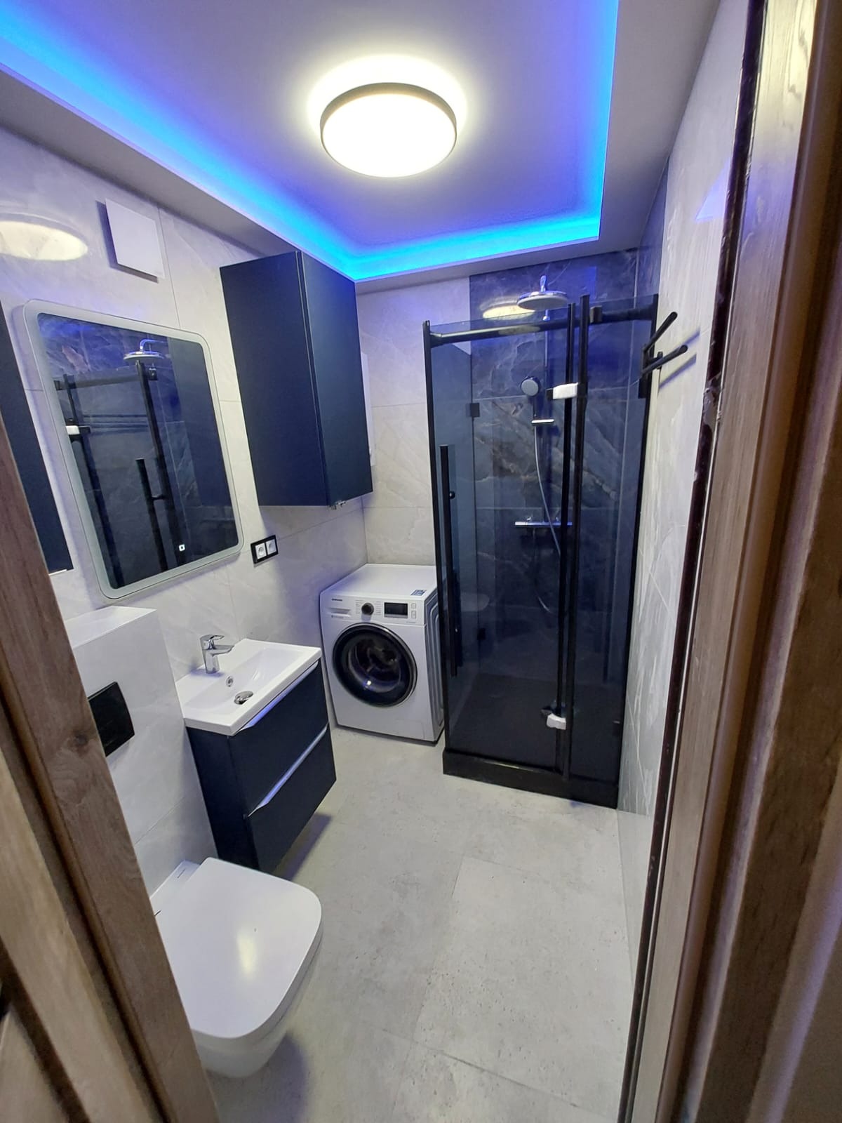 Nowoczesna łazienka z kabiną prysznicową typu walk-in, umywalką z szafką, toaletą i pralką. Podświetlenie LED na suficie. Minimalistyczny design.