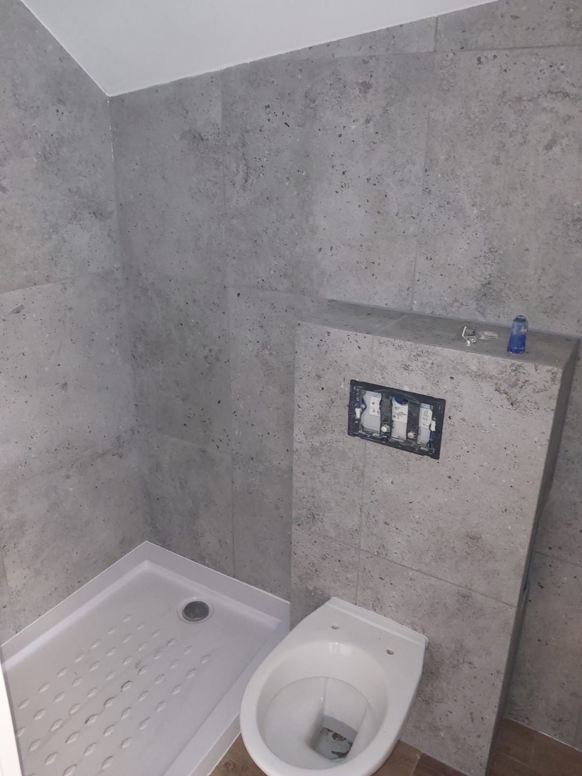 Wykończona łazienka z szarą, nakrapianą glazurą, białą kabiną prysznicową i podwieszaną toaletą. Widoczna instalacja podtynkową.