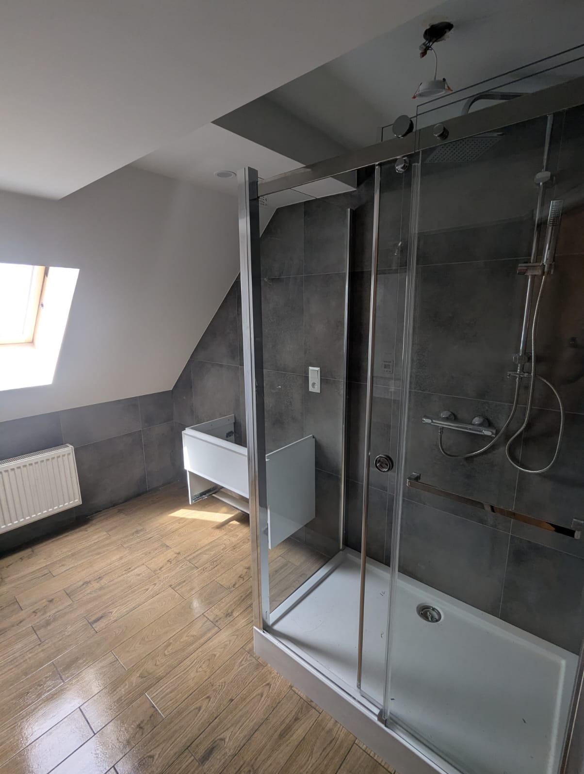 Nowoczesna łazienka na poddaszu z kabiną prysznicową, drewnopodobną podłogą i szarą glazurą. Umywalka częściowo zamontowana. Okno dachowe wpuszcza światło.
