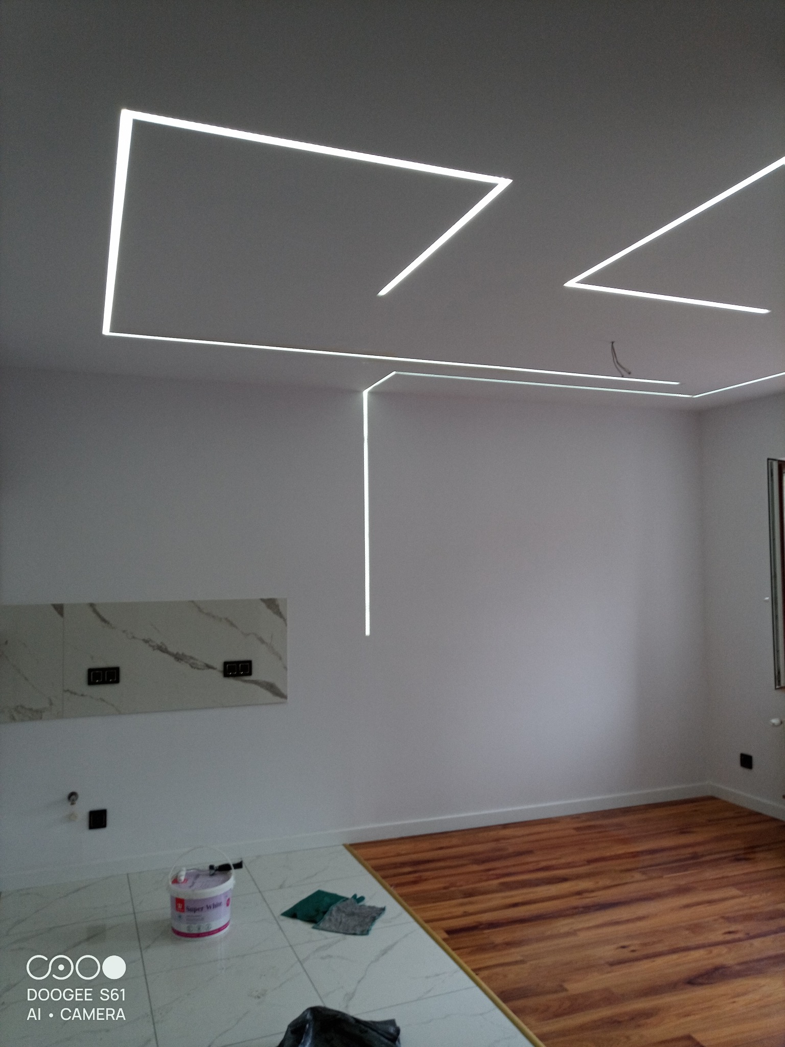 Nowoczesne oświetlenie LED w kształcie geometrycznym na suficie, białe ściany, marmurowe płytki podłogowe i drewniana podłoga. Widoczny fragment ściany z płytkami.