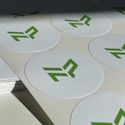 Cohera Studio - Arkusz białych, okrągłych naklejek z zielonym, nowoczesnym logo. Częściowo widoczny stos naklejek po lewej stronie. Jasne, naturalne oświetlenie.