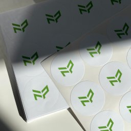 Cohera Studio - Stos białych, okrągłych naklejek z zielonym logo, ułożonych warstwami, oświetlonych naturalnym światłem, z widocznym cieniem.