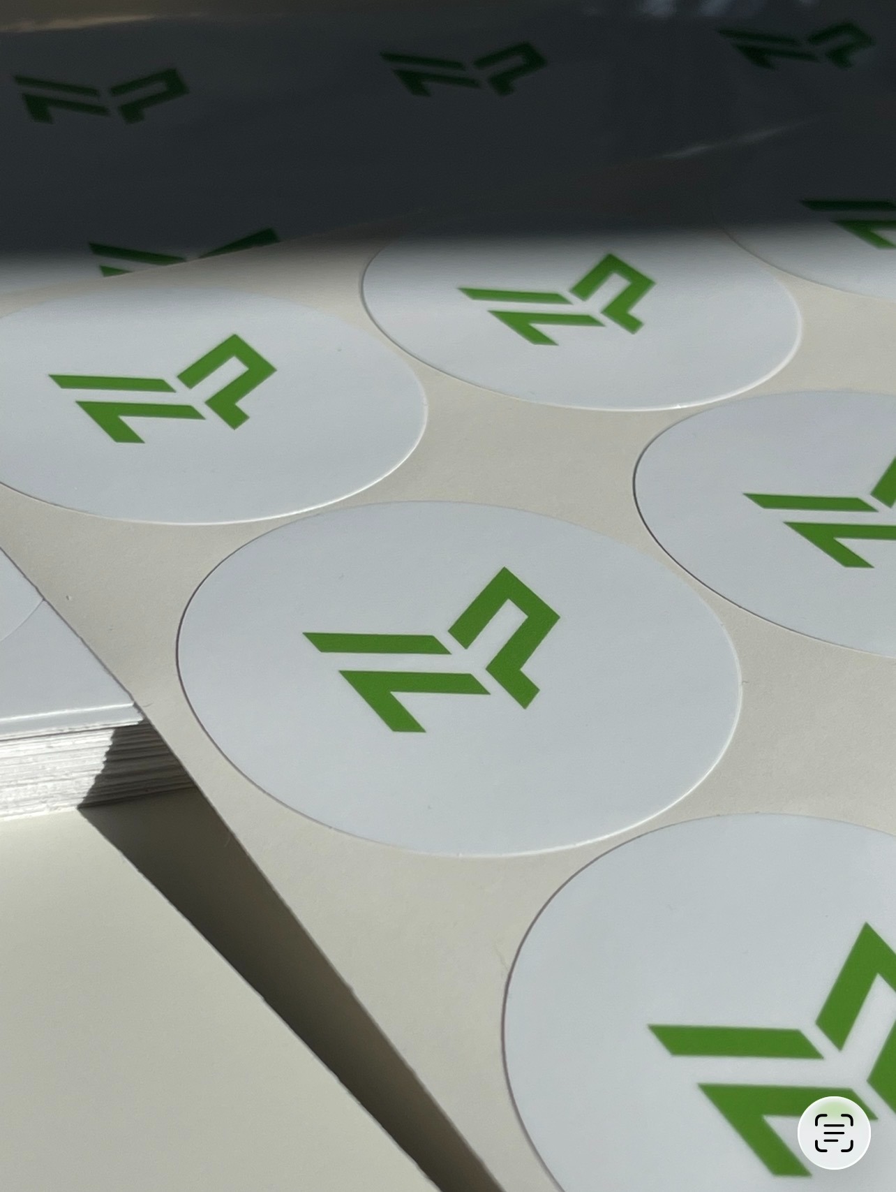 Arkusz białych, okrągłych naklejek z zielonym, nowoczesnym logo. Częściowo widoczny stos naklejek po lewej stronie. Jasne, naturalne oświetlenie.