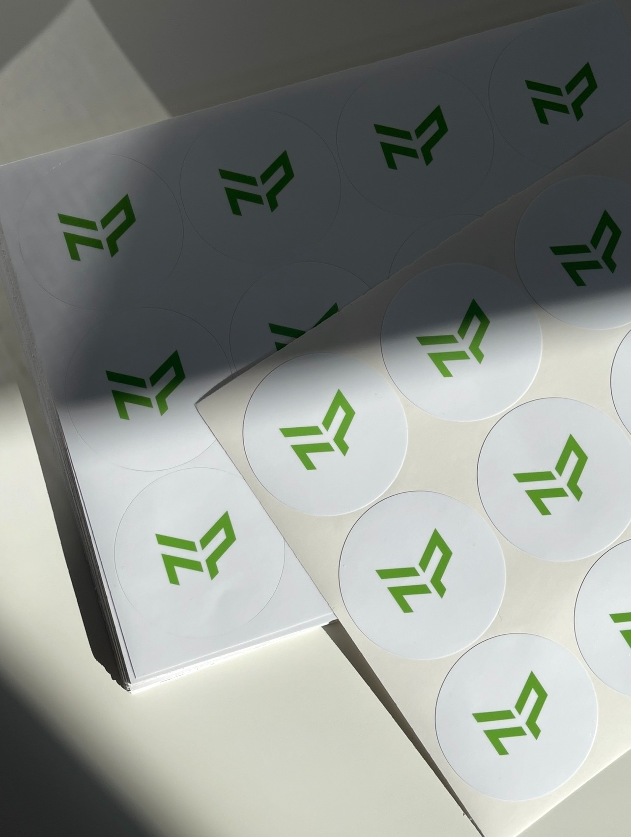 Stos białych, okrągłych naklejek z zielonym logo, ułożonych warstwami, oświetlonych naturalnym światłem, z widocznym cieniem.