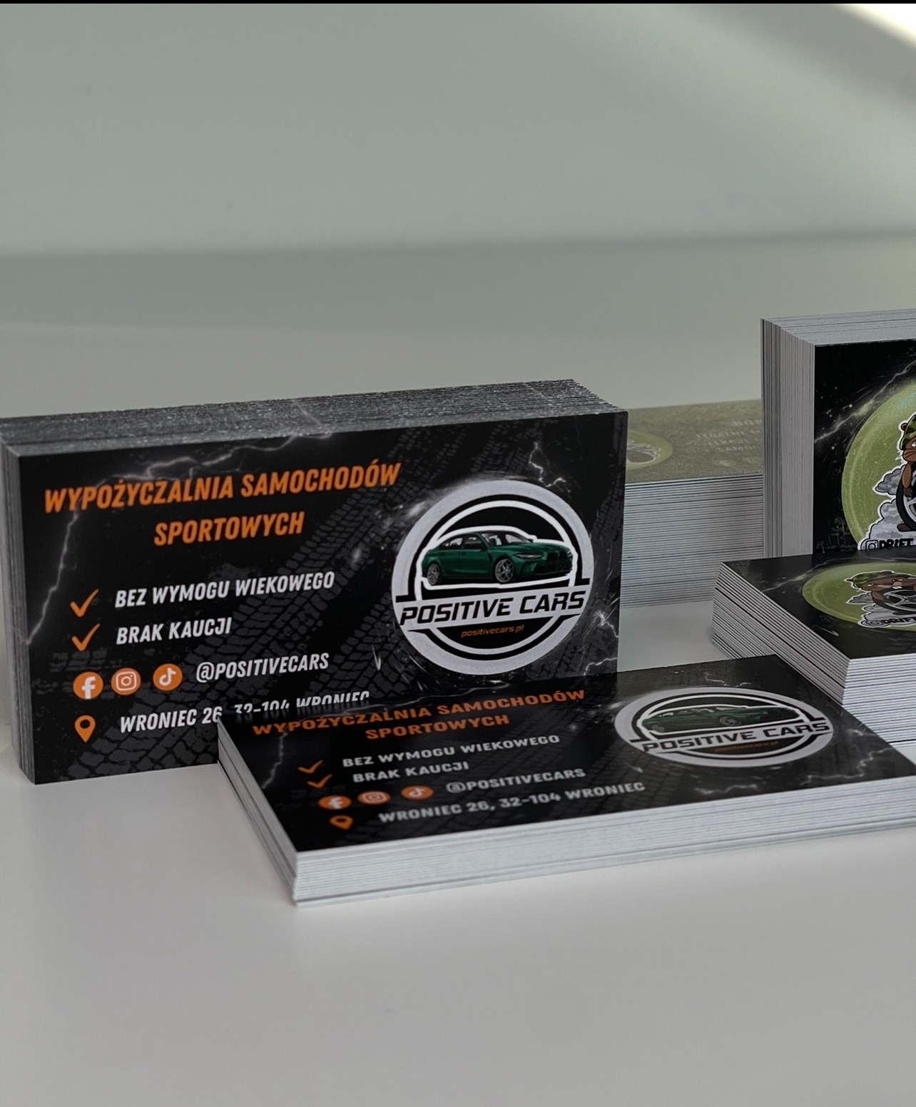 Stosy wizytówek z czarnym tłem, logo 'Positive Cars' i informacjami o wypożyczalni samochodów sportowych. Widoczne adresy social media i adres firmy.