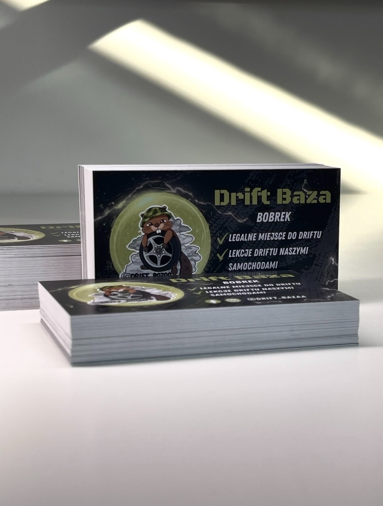 Stos wizytówek 'Drift Baza Bobrek' z ilustracją bobra w hełmie trzymającego oponę, na białym tle z refleksami światła. Widoczne hasła: legalne miejsce do driftu, lekcje driftu.