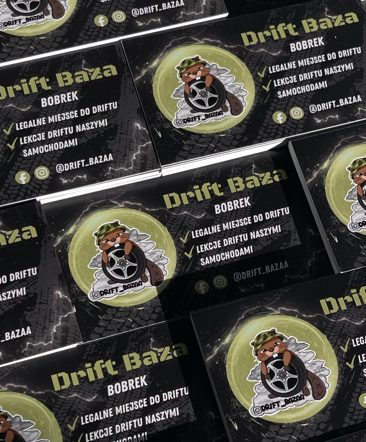 Stos wizytówek 'Drift Baza' z ilustracją bobra trzymającego oponę, na czarnym tle z motywem opon i błyskawic. Informacje o legalnym miejscu do driftu i lekcjach.