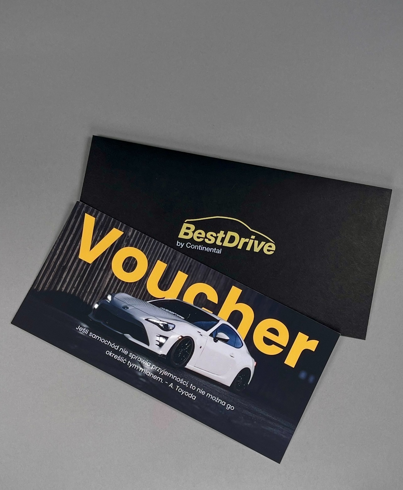 Czarno-złoty voucher BestDrive by Continental z białym samochodem sportowym, leżący na szarym tle obok czarnej koperty. Elegancki design, nowoczesny styl.