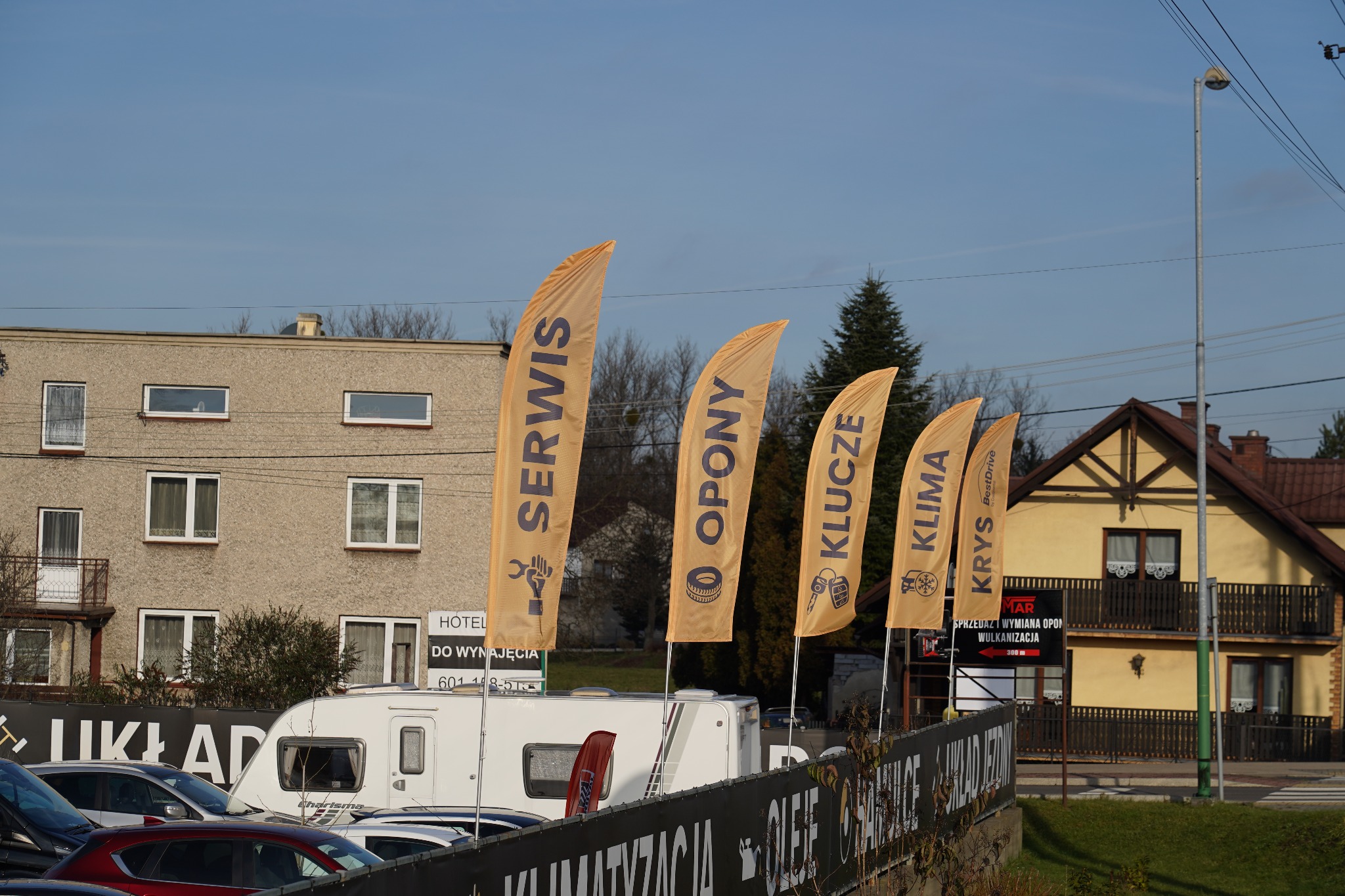 Żółte flagi reklamowe z napisami 'Serwis', 'Opony', 'Klucze', 'Klima' przed budynkami i samochodami. Widoczny szyld 'Sprzedaż i wymiana opon'.