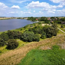 Snapium - Panoramiczny widok z lotu ptaka na jezioro, okoliczne pola, lasy i zabudowania wiejskie w słoneczny dzień. W tle widoczne turbiny wiatrowe.