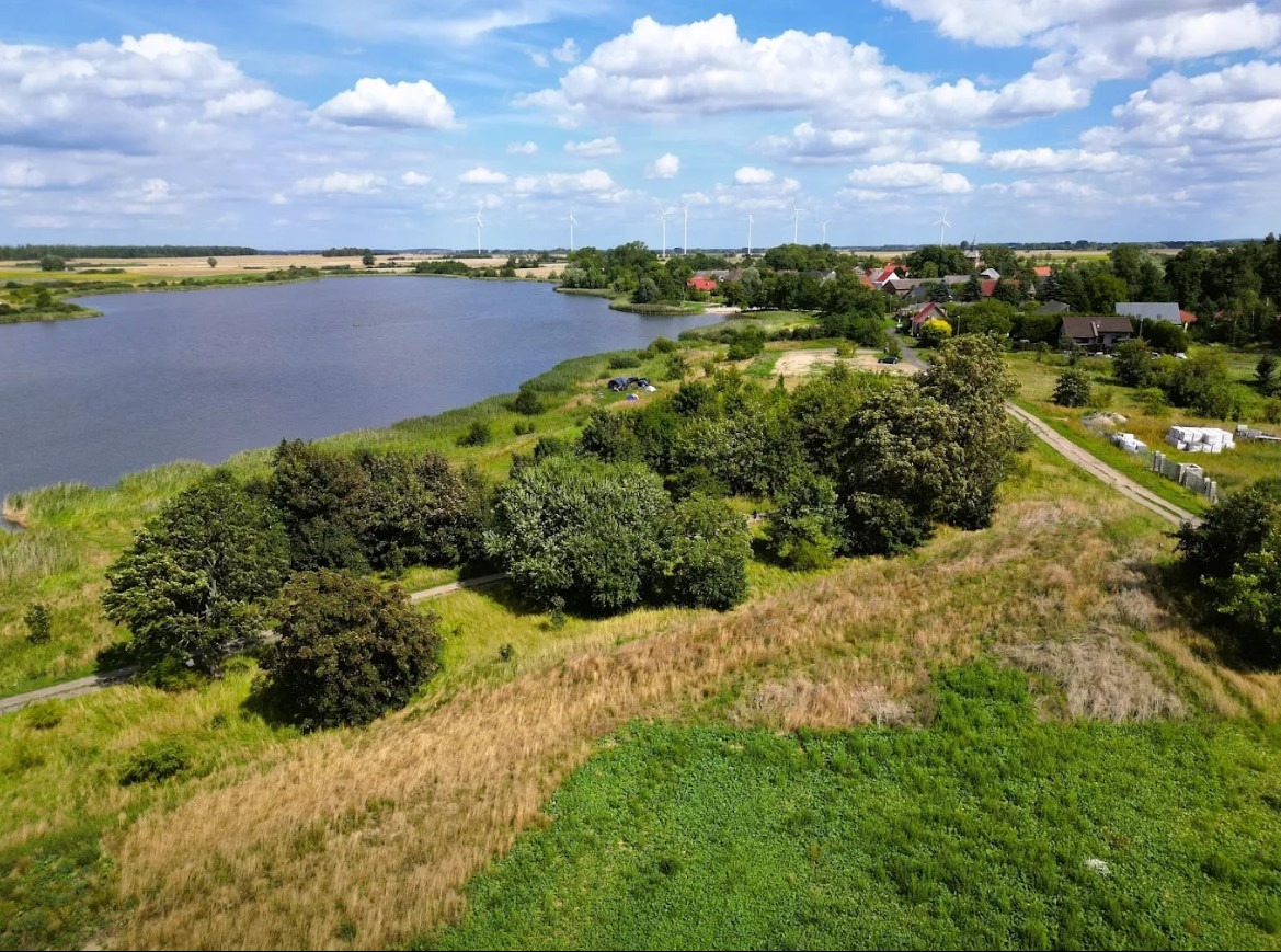 Panoramiczny widok z lotu ptaka na jezioro, okoliczne pola, lasy i zabudowania wiejskie w słoneczny dzień. W tle widoczne turbiny wiatrowe.