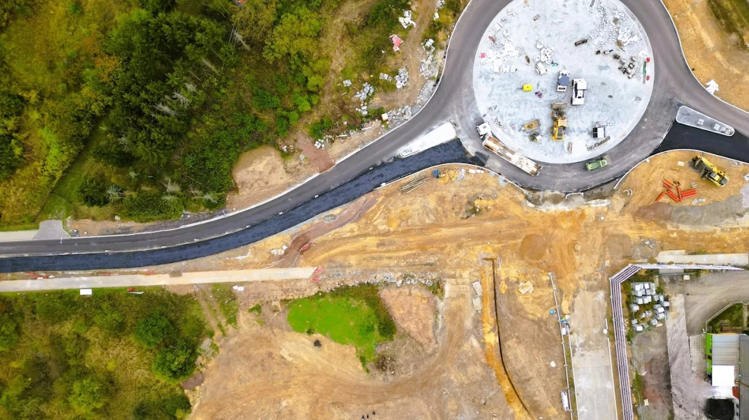 Widok z drona na budowę ronda z częściowo ukończoną nawierzchnią asfaltową, maszyny budowlane i teren przygotowany pod dalsze prace. Otoczenie to zieleń i zabudowania.