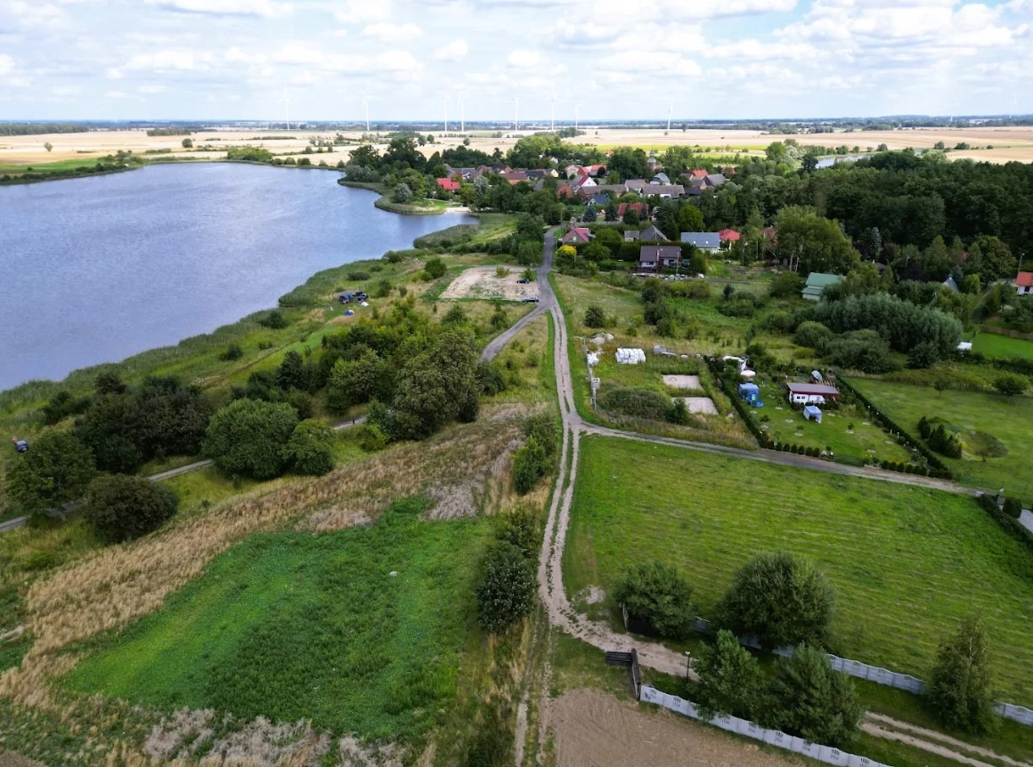 Panoramiczny widok z lotu ptaka na wieś z jeziorem, polami i turbinami wiatrowymi w tle. Widoczna droga gruntowa prowadząca do zabudowań.