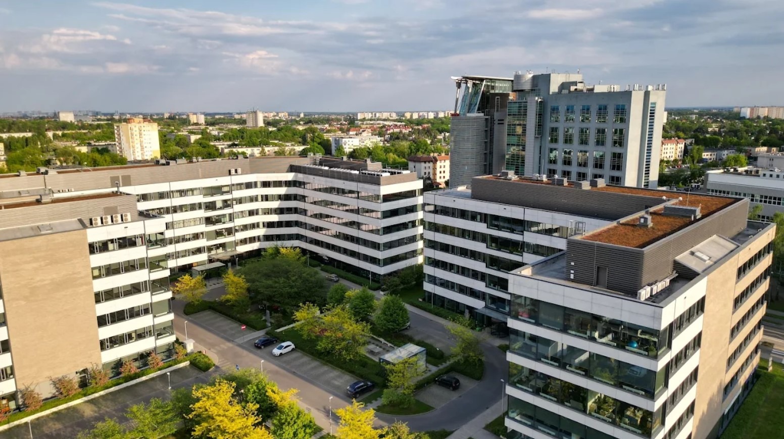 Kompleks biurowców z zielonymi dachami i parkingiem, widok z góry w słoneczny dzień. Nowoczesna architektura w otoczeniu zieleni miejskiej.