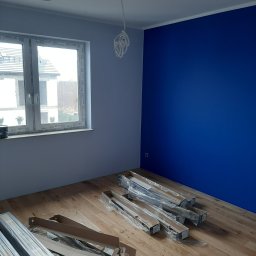 Oleksandr Partala - Pokój po malowaniu z niebieską ścianą i drewnianą podłogą. Na podłodze leżą panele podłogowe w opakowaniach, gotowe do montażu. Widoczne okno z widokiem na zewnątrz.