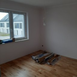 Oleksandr Partala - Jasna drewniana podłoga ułożona w pokoju z białymi ścianami. Przy oknie leżą materiały budowlane, a z sufitu zwisa kabel z żarówką. Widok na budynek za oknem.