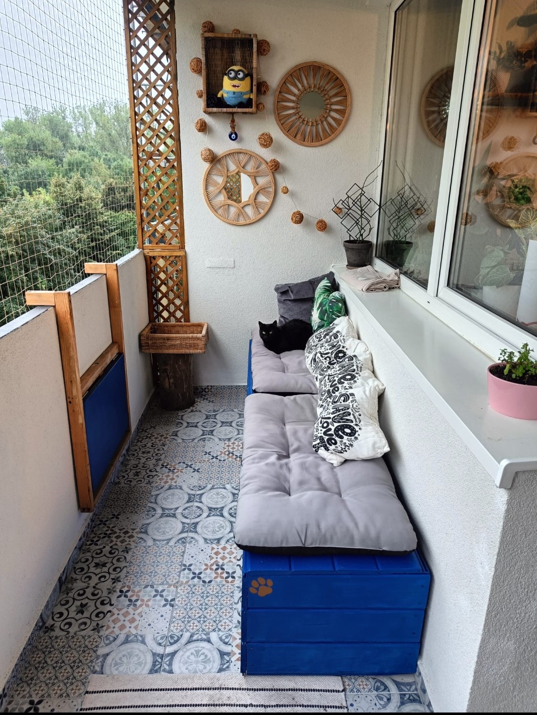 Przytulny balkon z wzorzystą podłogą, siedziskiem z szarymi poduchami i niebieską skrzynią z motywem łap. Na ścianie dekoracje i czarny kot.