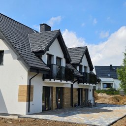 ARPOL-HOUSE INVESTMENTS SPÓŁKA Z OGRANICZONĄ ODPOWIEDZIALNOŚCIĄ - Nowoczesny dom z białymi ścianami i ciemną dachówką, z elementami dekoracyjnymi z drewna na elewacji. Widoczny fragment utwardzonego terenu przed budynkiem.