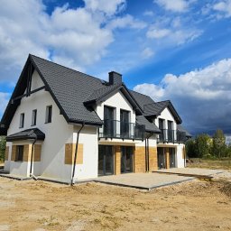 Arpol House - Nowoczesny dom z białymi ścianami i czarnym dachem, częściowo ocieplony, z widocznymi fragmentami elewacji w trakcie prac wykończeniowych. Błękitne niebo z chmurami w tle.