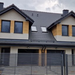 Arpol House - Nowoczesny dom z szarą dachówką i elementami drewnopodobnymi na elewacji, widoczny zza metalowego ogrodzenia. Uwagę zwracają okna dachowe i dwuspadowy dach.