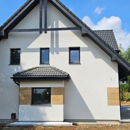 Arpol House - Nowoczesny dom z białą elewacją i czarnym dachem, widok z boku, z fragmentem podjazdu i zieleni. Na elewacji widoczne elementy dekoracyjne imitujące drewno.