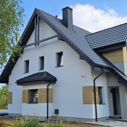 Arpol House - Nowoczesny dom z białą elewacją, czarnym dachem i elementami dekoracyjnymi z drewna. Widoczne okna, rynny i fragment ogrodu z drzewem. Ujęcie z boku, w słoneczny dzień.