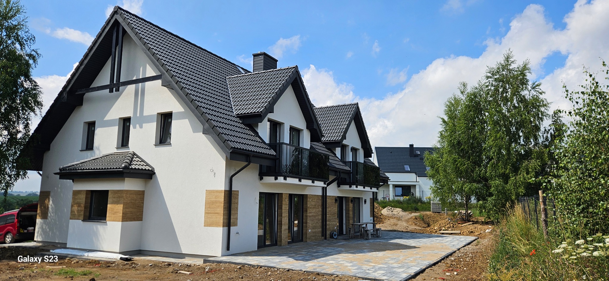 Nowoczesny dom z białymi ścianami i ciemną dachówką, z elementami dekoracyjnymi z drewna na elewacji. Widoczny fragment utwardzonego terenu przed budynkiem.