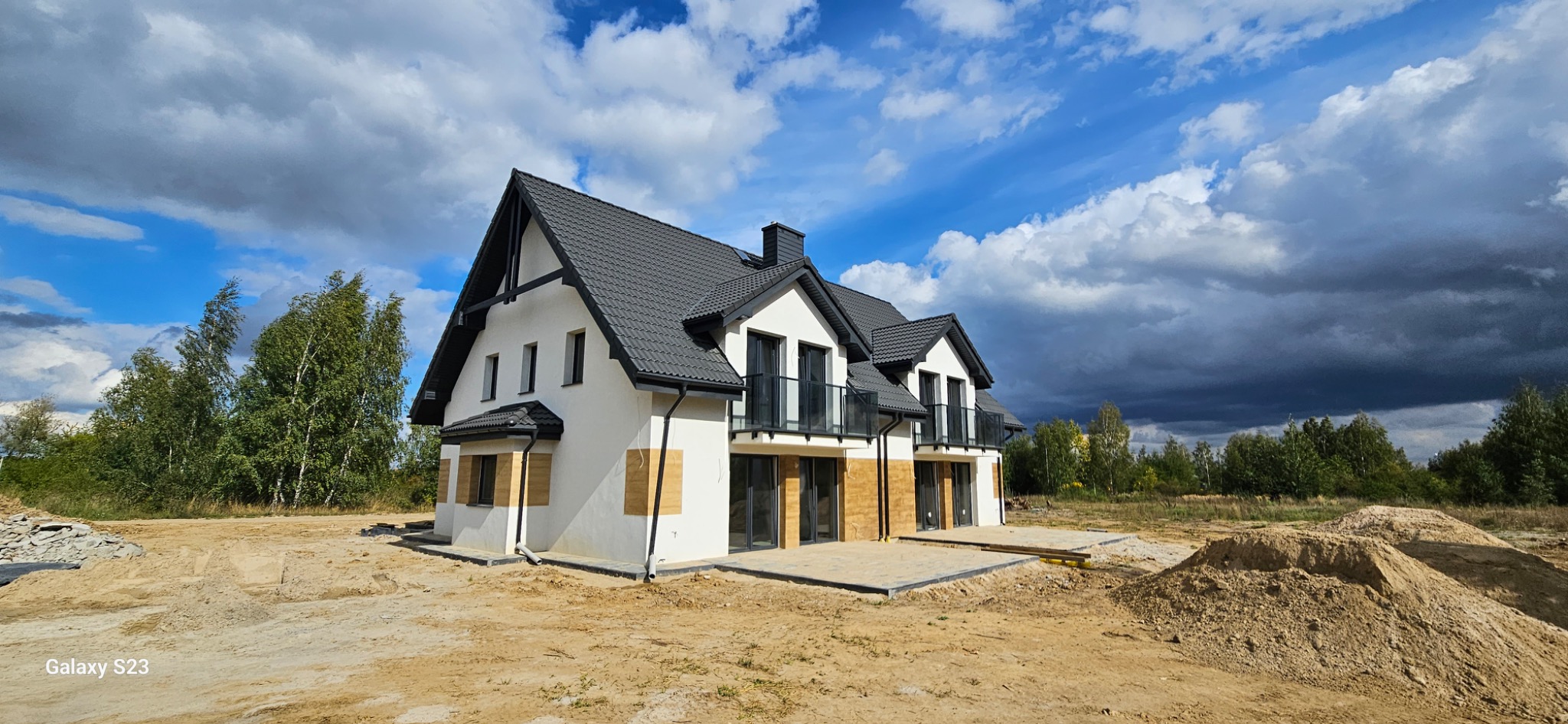 Nowoczesny dom z białymi ścianami i czarnym dachem, częściowo ocieplony, z widocznymi fragmentami elewacji w trakcie prac wykończeniowych. Błękitne niebo z chmurami w tle.